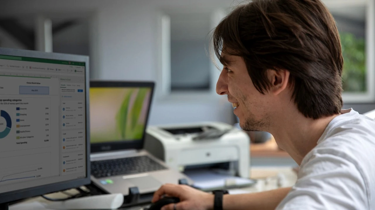 A young man smiles at a desktop computer in an office, with a laptop and a printer beside him
